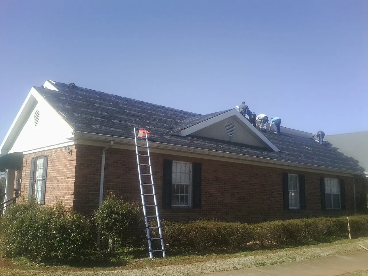 Crew tearing shingles off a brick ranch roof with a ladder set — Emergency Roof Repair work by our crew in Gardner