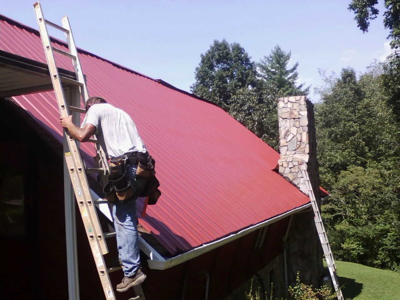 Roofer on a ladder beside a red metal roof and stone chimney — Soffit & Fascia Repair by Roofing Brothers in Gardner